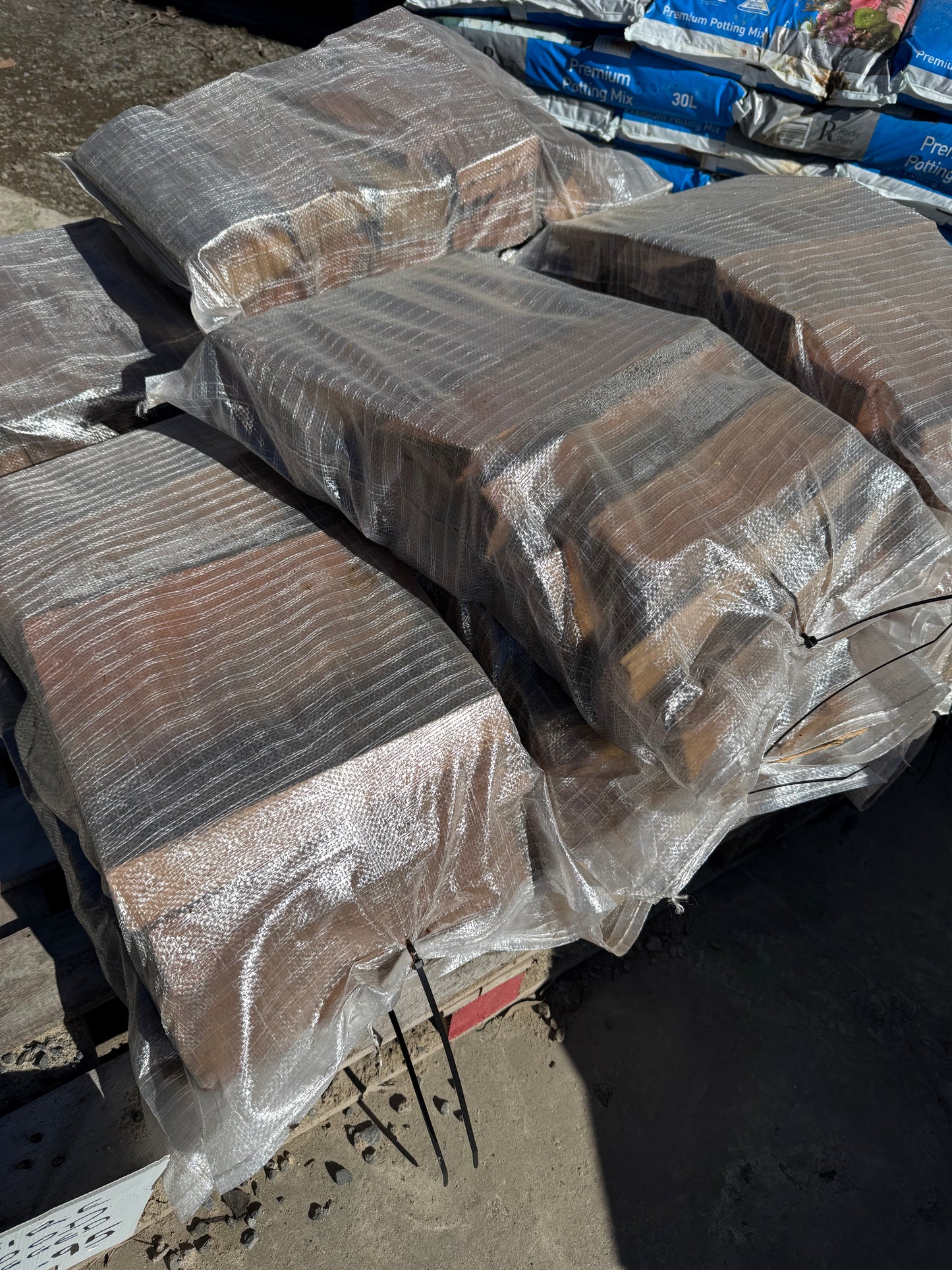 Bundles of FIRE WOOD TIMBER SPLIT are stacked outdoors, wrapped in clear plastic, with bags of landscaping material seen in the background.