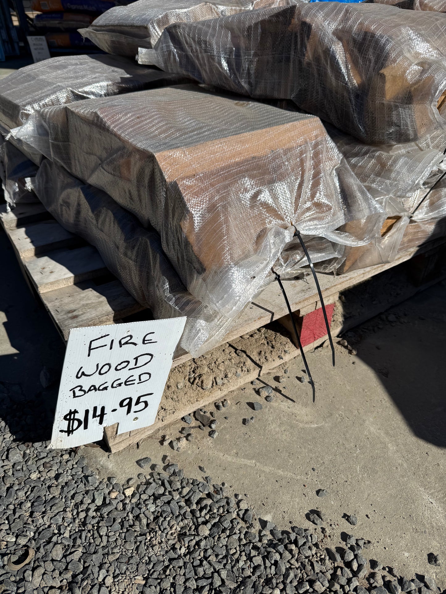 FIRE WOOD TIMBER SPLIT by 0 is stacked in plastic-wrapped bags on wooden pallets, with a handwritten sign reading FIRE WOOD BAGGED $14.95. The area features a gravel and dirt ground.