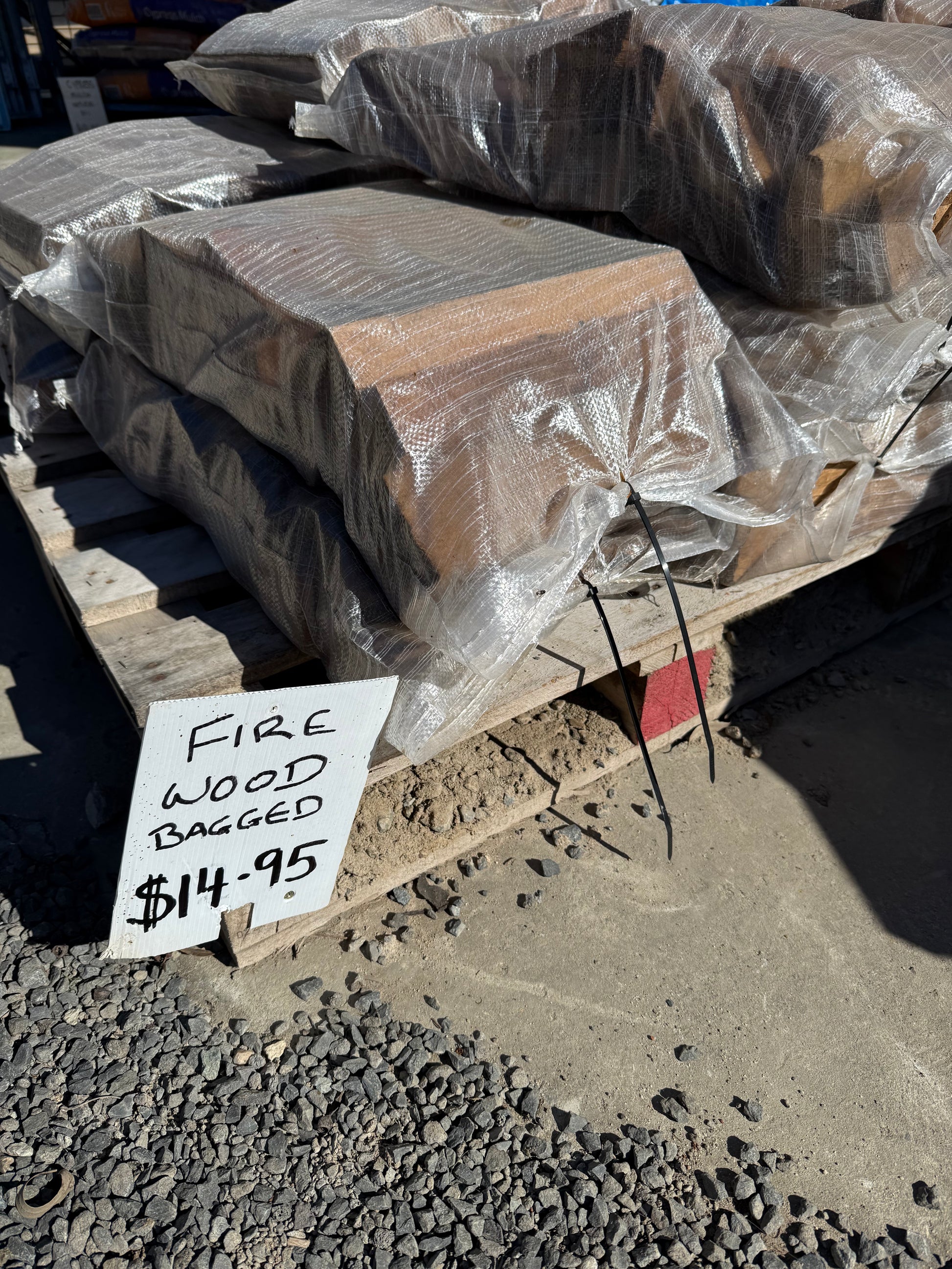 FIRE WOOD TIMBER SPLIT by 0 is stacked in plastic-wrapped bags on wooden pallets, with a handwritten sign reading FIRE WOOD BAGGED $14.95. The area features a gravel and dirt ground.