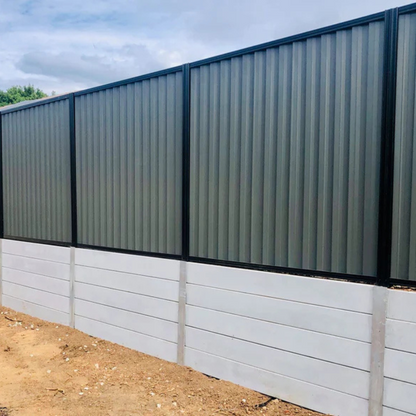 A tall fence with upper gray corrugated metal panels in black frames and a lower section made of Smooth Plain Grey 2 bar Concrete Sleeper stands on bare soil under a partly cloudy sky.
